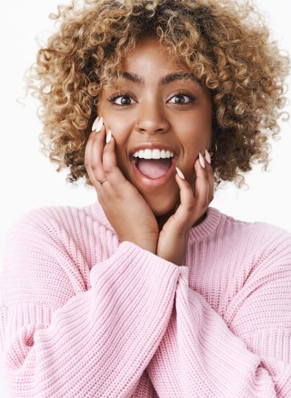 Studio shot of amused and excited good-looking african female with fair curly hair smiling and dropping jaw from amazement holding hands on cheeks standing thrilled over white background. Copy space